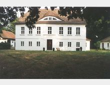 Weiße, zweistöckige Villa mit symmetrischer Fensteranordnung und einem Satteldach, umgeben von einer Wiese und Bäumen. Ein zentral platzierter Holz-Eingang ist von kleinen Büschen flankiert, und Nebengebäude sind teilweise sichtbar.