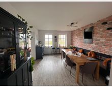 Esszimmer mit hellem Boden, einer Ziegelwand auf der rechten Seite und einem großen Holztisch, der von Stühlen und einer gepolsterten Bank mit orangefarbenen Kissen umgeben ist. Links befindet sich ein schwarzer Glasschrank mit Dekoration, während zwei Fenster den Raum erhellen.