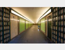 Langer Unterführungsgang mit beigefarbenen Fliesenwänden und grünen geschlossenen Rolläden auf beiden Seiten, darüber durchgehende Beleuchtung an der Decke. Metallgittertüren befinden sich an den Eingängen zu beiden Seiten.