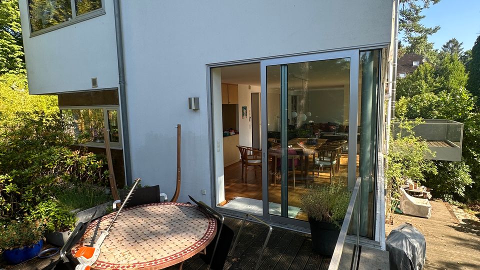 Moderne Hausfassade mit großer Schiebetür aus Glas, die auf eine Holzterrasse führt. Auf der Terrasse stehen ein runder Metalltisch, Stühle und Pflanzkübel; seitlich verläuft ein Glasgeländer mit Blick ins grüne, baumbestandene Umfeld.