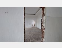 Ein leerer Innenraum mit weißen Wänden und mehreren offenen Türdurchbrüchen, die teilweise unverputzt sind und Ziegel freilegen. Der Boden ist roh und wirkt unbehandelt, während durch die hinteren Öffnungen Tageslicht einfällt.