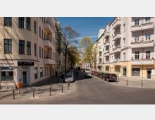 Straßenszene in einer Wohngegend mit mehrstöckigen Altbauten, Balkonen und pastellfarbenen Fassaden. Entlang der Straße stehen geparkte Autos, vereinzelte Bäume spenden Schatten, und im Vordergrund liegt ein Café an der Ecke.