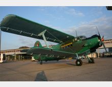Ein grünes Doppeldecker-Flugzeug mit der Aufschrift "Classic Aircraft" steht auf einem offenen Flugfeld. Im Hintergrund sind ein Gebäude mit großen Fenstern und ein Bus zu sehen, bei klarem Himmel und tief stehender Sonne.