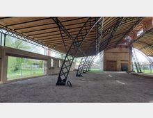 Teilweise offene Industriehalle mit gewölbtem Holzdach auf schwarzen Metallträgern, umgeben von einer alten Backsteinstruktur. Im Hintergrund sind Bäume, ein Zaun und weitere Gebäude sichtbar.