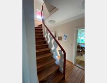 Holzinnentreppe mit geschwungenem weißen Geländer und rotem Handlauf in einem Wohnraum. Im Hintergrund sind eine Holztür zum Essbereich und Wanddekoration sichtbar.