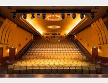 Innenansicht eines Theatersaals mit ansteigenden Sitzreihen in der Mitte und seitlichen Treppen mit Stufenbeleuchtung. An der Decke hängen zwei große Kronleuchter sowie eine Licht- und Techniktraverse; die Wände sind holzverkleidet und von Wandleuchten gerahmt.