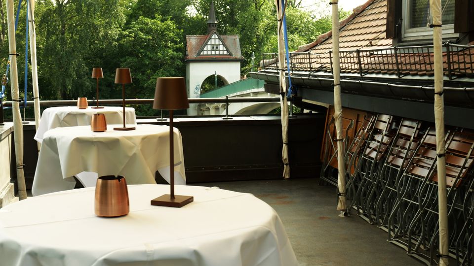 Überdachte Restaurantterrasse mit runden Tischen in weißen Tischdecken und schlanken Tischlampen; seitlich stehen zusammengeklappte Stühle an einer Hauswand mit Ziegeldach. Im Hintergrund sind Bäume sowie ein kleiner Turm- und Torbogenbau neben einer Brücke zu sehen.