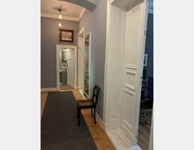 Langer Flur in einer Wohnung mit grauen Wänden, Holzdielenboden und dunklem Läuferteppich, beleuchtet von hängenden Glühbirnen. Weiße Zimmertüren und Türrahmen, gerahmte Bilder an der Wand sowie ein Stuhl neben einer Vitrine; am Ende ist ein kleines Bad sichtbar.