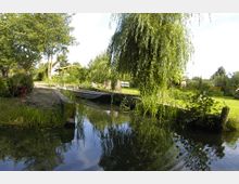 Eine grüne Gartenlandschaft mit einem kleinen Bach, der von einer Holzbrücke überspannt wird. Im Hintergrund sind eine Weide, andere Bäume, eine Wiese sowie eine Sitzbank sichtbar.