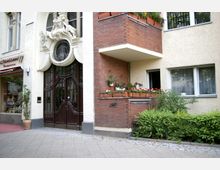 Fassade eines Wohnhauses mit verziertem dunkelbraunem Holzeingang, reichhaltigen Stuckarbeiten und einem kunstvollen Rundfenster darüber. Daneben ein Balkon mit roten Ziegeln, bepflanzt mit Blumen in Blumenkästen, sowie ein Geschäft mit einer Markise.