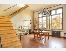 Wohnraum mit hellen Holzelementen, einer offenen Holztreppe, hohen Fenstern und einem Kronleuchter. Im Raum stehen ein Esstisch mit Stühlen, ein Regal mit Geschirr sowie eine Leiter an der Wand.