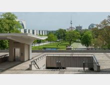 Städtische Umgebung mit einem gepflasterten Platz, einer überdachten Aufzugsanlage und einem Treppenabgang, umgeben von grünen Bäumen und gepflegten Hecken. Im Hintergrund sind moderne Regierungsgebäude, die Kuppel des Reichstags und der Berliner Fernsehturm zu sehen.