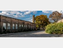 Ein historisches Backsteingebäude mit großen, bogenförmigen Fenstern steht an einer mit Kopfsteinpflaster bedeckten Straße. Im Hintergrund ist ein hoher Schornstein sichtbar, umgeben von Bäumen mit herbstlichem Laub und dichtem grünen Gebüsch.
