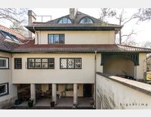 Zweistöckiges Wohnhaus mit hellbeiger Fassade und roten Ziegeldächern, verziert mit Gitterfenstern und einem kleinen Vorbau, der von Säulen getragen wird. Der Innenhofbereich ist teilweise überdacht, und im Hintergrund sind Bäume erkennbar.