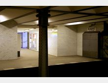 U-Bahn-Station mit weiß gefliesten Wänden, einer ausgeschilderten Ausganganzeige und Werbeplakaten an der Wand. Im Vordergrund eine Stahlstütze und ein Bahnsteigabschnitt.