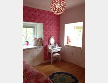 Ein helles Kinderzimmer mit rosa Tapeten und weißen Möbeln. Im Raum befinden sich ein Frisiertisch mit Hocker, eingebauter Stauraum unter zwei Fenstern und eine leuchtende, dekorative Kugellampe an der Decke.