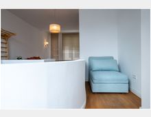 Helles Wohnzimmer mit weiß gestrichenen Wänden, Holzfußboden und einer Stufe mit geschwungener Abtrennung. Auf der rechten Seite steht eine hellblaue Liegesitzfläche, im Hintergrund befindet sich eine Wand mit Jalousien vor den Fenstern sowie eine Deckenleuchte.