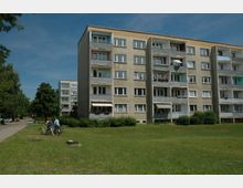 Mehrstöckige Wohnblöcke mit Balkonen und teilweise geschlossenen Markisen, umgeben von einer Grünfläche und Bäumen; im Vordergrund zwei Personen mit Fahrrädern.