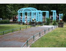 Ein kleiner Park mit einer hellblauen, gitterartigen Pergolakonstruktion im Zentrum, umgeben von Rasen und Bäumen. Im Vordergrund führt ein gepflasterter Weg mit Geländern zu der Pergola, die auf Steinsockeln mit goldenen Kugeln an den Ecken steht.