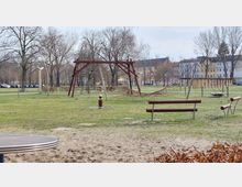 Ein offener Spielplatz mit Klettergerüst aus Holz und Seilen in einer Grünfläche mit Bäumen im Hintergrund. Im Vordergrund sind Bänke und ein Sandstreifen, im Hintergrund sind geparkte Autos und mehrstöckige Wohngebäude zu sehen.