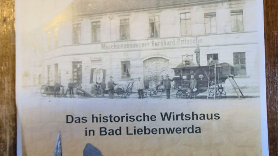 Flyer auf einer Holzoberfläche mit der Aufschrift „Alte Schmiede“ und einem historischen Foto eines zweigeschossigen Gasthauses. Darunter steht „Das historische Wirtshaus in Bad Liebenwerda“ sowie eine Telefon­nummer.