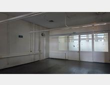 Leerstehender Raum mit weißen Wänden und dunklem Boden. Die Rückwand besteht aus einer Glasfront mit teils mattierten Scheiben, durch die ein Korridor sichtbar ist; an der linken Wand befinden sich Rohre, eine Uhr, Steckdosen und eine Tür.