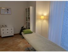 Ein heller Raum mit einer Behandlungsliege, einem grünen Kissen darauf, einer weißen Kommode mit kleinen Schubladen und einem Schrank mit Glastüren. Eine Stehlampe spendet warmes Licht, und ein Vorhang sowie eine Wanddekoration runden den Raum ab.