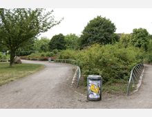 Ein Park mit geschwungenen, unbefestigten Wegen, umgeben von Bäumen, Büschen und Grünflächen. Im Vordergrund steht ein Abfalleimer mit einem Plakat, und Metallgeländer säumen die Wege.