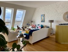 Wohnzimmer unter einem Dachfenster mit schrägen, großflächigen Fenstern, die auf eine städtische Umgebung blicken. Der Raum ist eingerichtet mit weißen Sofas, einem runden Beistelltisch, dekorativen Pflanzen, einer gemauerten weißen Ziegelwand, einer Wanddekoration und einem goldenen Sideboard.