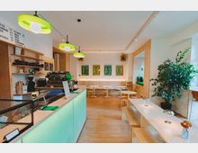 Modern café interior with a mint-green service counter, open wood shelving, and a seating area of small round tables and long benches on light wood floors. Green pendant lights hang from the ceiling, colorful posters line the back wall, and a large potted plant sits near a windowed nook.