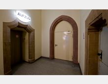 Flur mit gelblicher Wandfarbe und Türrahmen aus rötlichem Sandstein. Links befindet sich eine Aufzugsöffnung mit einem beleuchteten Schild "Aufzug", rechts eine cremefarbene Tür mit einem Spitzbogenelement.