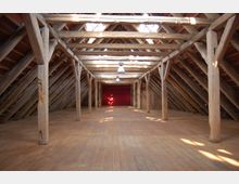 Dachboden eines historischen Gebäudes mit sichtbarem Holzbalkengerüst und Holzdielenboden, beleuchtet durch Lichtstrahlen, die durch Dachfenster fallen; am Ende des Raums befindet sich eine rote Wand.