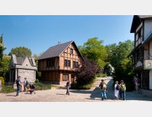 Ein kleiner Platz mit Kopfsteinpflaster, umgeben von Fachwerkhäusern und einem steinernen Gebäude mit Spitzdach. Im Hintergrund ist ein grüner Park mit einer Statue zu sehen, während mehrere Menschen über den Platz laufen oder stehen.
