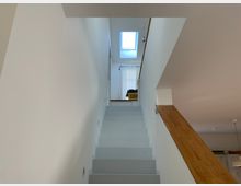 Weiße Innentreppe mit einem Holzgeländer auf der rechten Seite, die zu einem oberen Stockwerk führt. Oben befinden sich ein großes Fenster mit Sicht nach draußen und ein Vorhang, der Tageslicht einfängt.