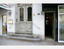 Fassade eines Altbaus mit heller Putzfassade, einer großen verzierten Doppeltür aus Holz mit Glasfenstern und drei Steinstufen, die zum Eingang führen. Rechts befindet sich ein schmaler Durchgang mit Hausnummer 51, daneben ein Teil einer Geschäftsfront mit einem Ladenfenster und Schild.