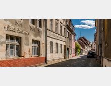 Schmale gepflasterte Gasse in einer Kleinstadt, gesäumt von teils renovierungsbedürftigen, historischen Gebäuden mit Putz- und Ziegelwänden. Im Hintergrund sind weitere Häuser mit roten Ziegeldächern sowie ein blauer Himmel mit weißen Wolken zu sehen.