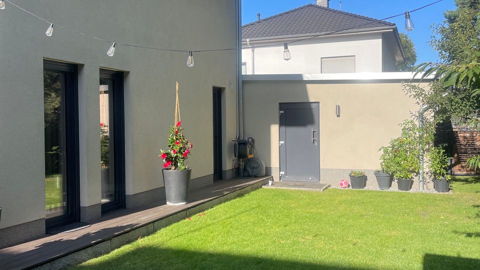 Moderne Hausfassade mit beigem Putz und großen schwarzen Fenstern, die zu einer Holzterrasse führen. Vor dem Haus befindet sich ein gepflegter Rasen, umgeben von vereinzelten Pflanzen in Töpfen und Bäumen im Hintergrund.