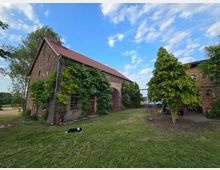Backsteinbauernhof mit rotem Ziegeldach, teilweise von grünen Kletterpflanzen bedeckt, daneben ein kleiner Innenhof mit Bäumen und Wiese. Im Vordergrund liegt eine schwarz-weiße Katze auf dem Rasen, im Hintergrund ist freies Feld zu sehen.