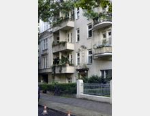 Mehrstöckiges Wohnhaus im Altbaustil mit verzierten Fassadenelementen und mehreren Balkonen, die teilweise mit Pflanzen dekoriert sind. Im Vordergrund befinden sich ein Gehweg, eine niedrige Mauer mit Zaun sowie ein Fahrrad.