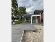 Ein langer überdachter Holzsteg führt vom rechten Rand eines Backsteingebäudes mit dunkelgrauem Dach entlang einer Wiese und eines sandigen Bereichs. Im Hintergrund sind Bäume und ein Fluss zu sehen, unter einem bewölkten Himmel.