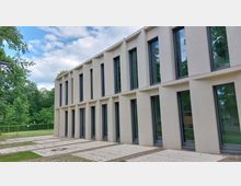 Moderne Gebäude-Fassade aus hellem Putz mit großen, schmalen Fenstern in symmetrischer Anordnung. Im Vordergrund gepflasterte Wege, dahinter eine Grünfläche mit Bäumen und Sträuchern.