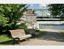 Zwei Holzbänke auf einem gepflasterten Weg in einem parkähnlichen Umfeld nahe einem Flussufer. Im Hintergrund befindet sich eine steinerne Treppe, eine Brücke aus grünem Metall und dichtes Blattwerk, das den Bereich umrahmt.