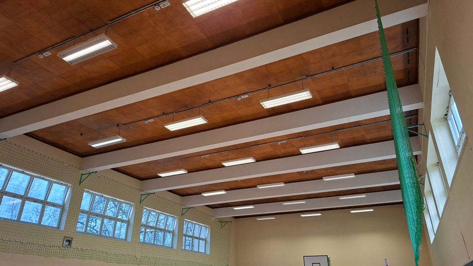 Blick in eine Sporthalle mit hoher Holzdecke, weißen Querträgern und langen Deckenleuchten. An den Wänden befinden sich große Fenster sowie ein gespanntes Schutznetz, daneben hängt ein grünes Seitennetz.