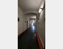 Langer, schmaler Gebäudeflur mit heller Decke, dunklem Boden und roten Bodenleisten. Beidseitig hängen Bilder an den Wänden, und am Ende des Flurs ist eine Glastür mit Blick auf Tageslicht erkennbar.