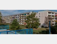 Mehrstöckiger Wohnblock mit einer langen Reihe von kleinen Balkonen, daneben mehrere Bäume und eine Grünfläche. Im Vordergrund ist ein blaues Geländer zu sehen, das zu einer Treppenabgang führt.