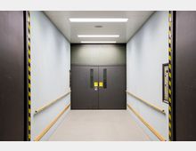 Ein leerer, rechteckiger Lastenaufzug mit glatten, hellblauen Wänden, gelben Handläufen an beiden Seiten und schwarzen Schiebetüren mit kleinen Fensteröffnungen. Der Boden ist grau, und die Decke enthält mehrere rechteckige Lichtpaneele.