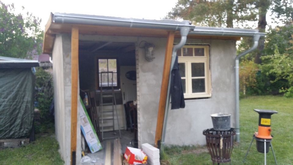 Kleines Gartenhaus aus grauem Putz mit flachem Dach, Regenrinnen und einem offenen Anbau mit Holzstützen; im Inneren sind Leitern und Baumaterial zu sehen. Daneben befindet sich ein Fenster mit hellem Rahmen, umgeben von Rasen und Bäumen.