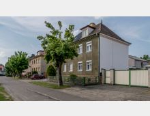 Wohnstraße mit zweistöckigem Reihenhaus im Vordergrund, das eine grün-braune Fassade und weiße Fenster hat. Vor dem Gebäude stehen ein kleiner Garten mit Zaun, ein gepflasterter Gehweg sowie ein Baum; daneben befindet sich eine Einfahrt mit einem hellen Tor.