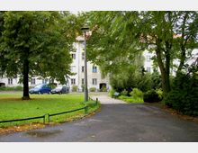 Grüne Parkanlage mit gepflastertem Weg und Bäumen, im Hintergrund ein mehrstöckiges Wohngebäude mit weißer Fassade und roten Dachziegeln. Im Vordergrund befinden sich eine Laterne, geparkte Autos sowie eine Bank am Wegesrand.