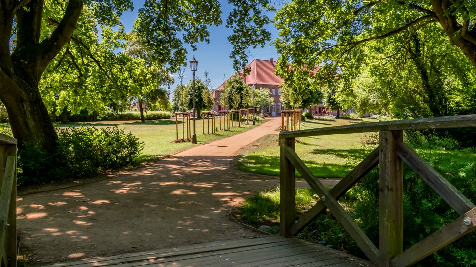 Blick von einer kleinen Holzbrücke auf einen geschwungenen Sandweg durch einen grünen Park mit großen, schattenspendenden Bäumen und Rasenflächen. Im Hintergrund steht ein Gebäude mit rotem Ziegeldach, flankiert von jungen Bäumen und einer Laterne entlang des Weges.